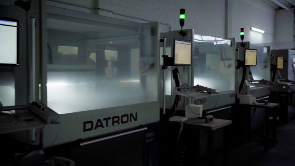 Reihe von DATRON-CNC-Maschinen in einer dunklen Werkhalle, jeweils mit Monitor, durchsichtiger Schutzscheibe und grüner Signalleuchte.
