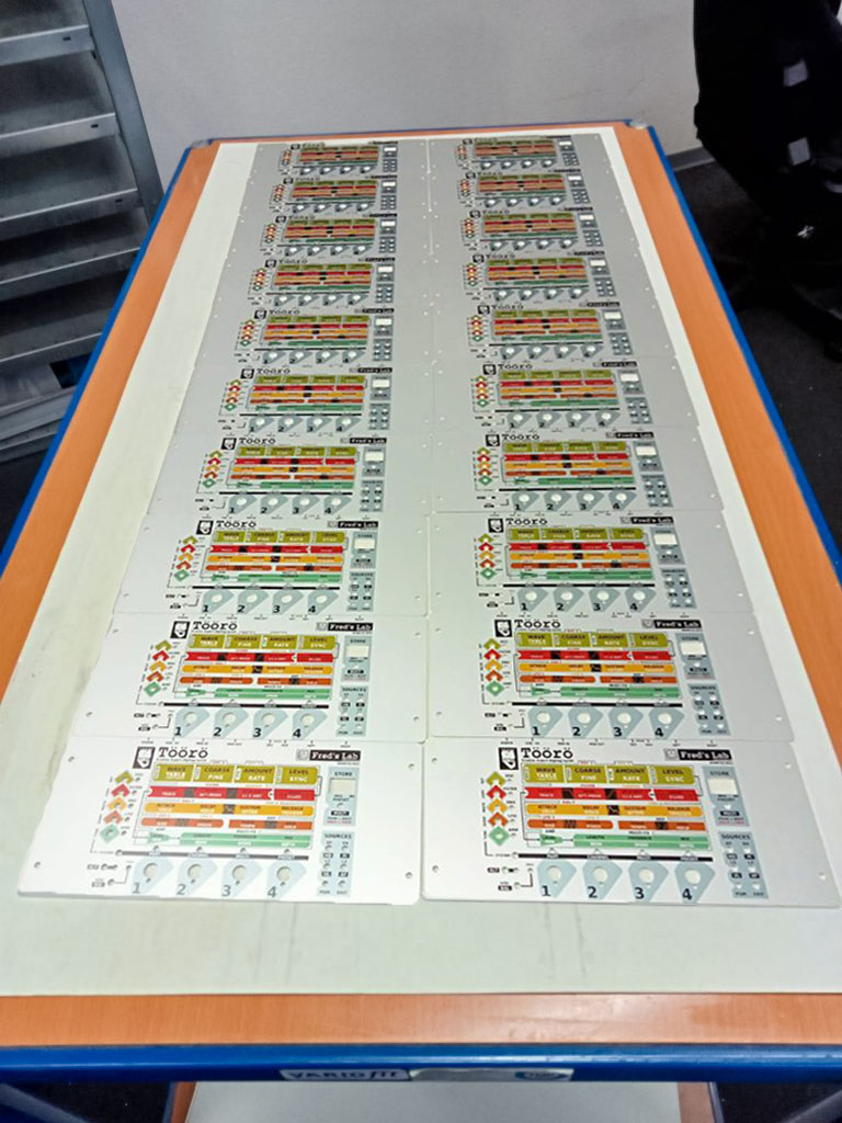 Mehrere weiße Frontplatten für Elektronikgeräte mit farbigen Beschriftungsfeldern und gedruckten Drehknopfsymbolen in zwei Spalten gleichmäßig auf einem Werkbanktisch angeordnet, im Hintergrund Werkstattregal und Boden sichtbar.