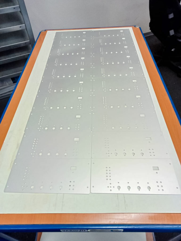 Mehrere hellgraue rechteckige Metallplatten mit Reihen gebohrter Löcher und rechteckigen Aussparungen, ordentlich in mehreren gleichmäßigen Reihen auf einer langen orangenen Tischfläche mit blauer Kante in einer Werkstattumgebung angeordnet.